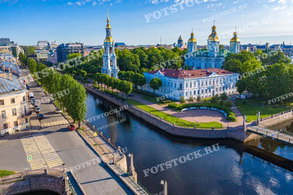 Это снимок с воздуха Никольского морского собора в Санкт-Петербурге, расположенного на пересечении Крюкова канала и канала Грибоедова. Солнце мягко освещает здание и золоченые купола, отражаясь в спокойных водах каналов. Зеленые аллеи и спокойные улицы придают сцене ощущение умиротворения. Фотография идеально подходит для путеводителей, исторических книг о Санкт-Петербурге, а также для любого контента, посвященного российской культуре и архитектуре.